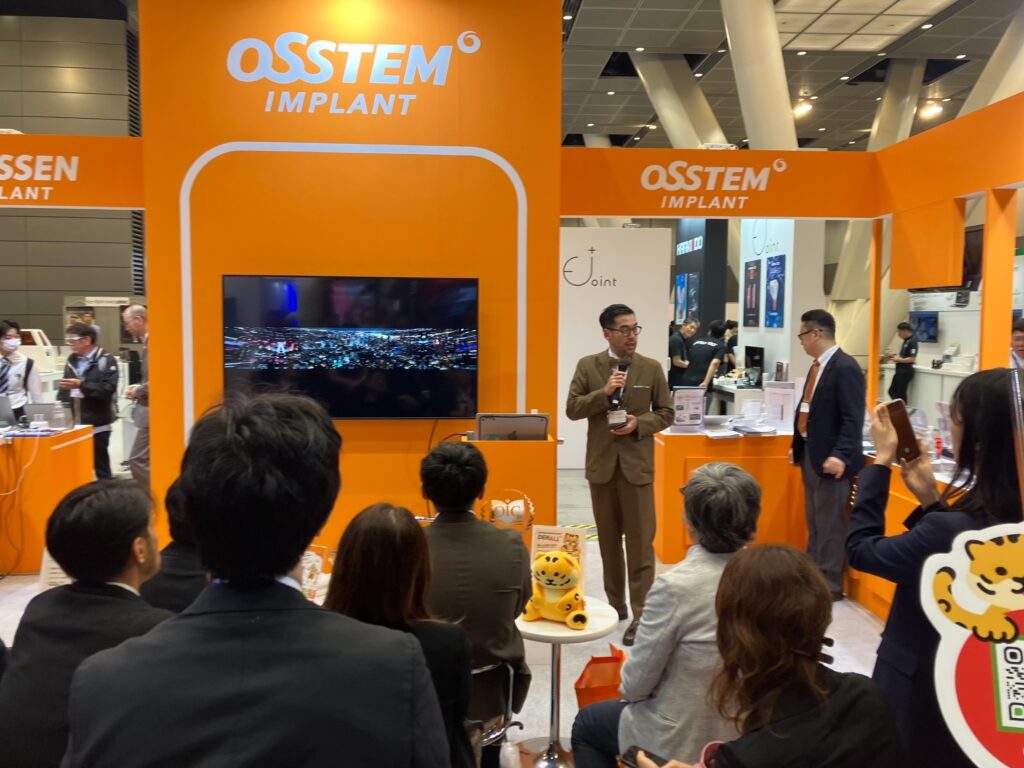 OSSTEM JAPANブースのモニター横で橋口隼人がレクチャーを行い、参加者が聴講している様子
